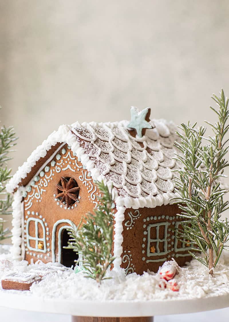 A frosted gingerbread house on a cake platter. - decorated gingerbread houses, royal icing, parchment paper