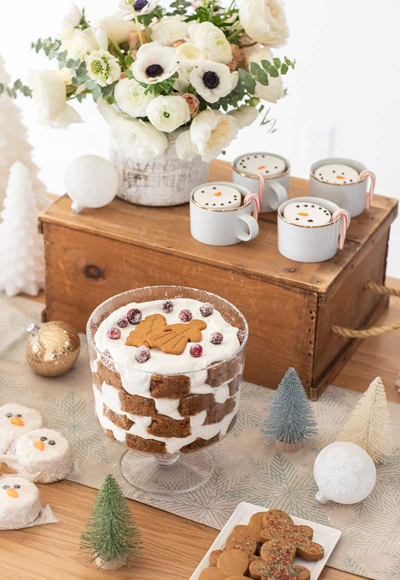 Christmas dessert table with white flowers, gold cups with marshmallow snowmen, gingerbread trifle. 