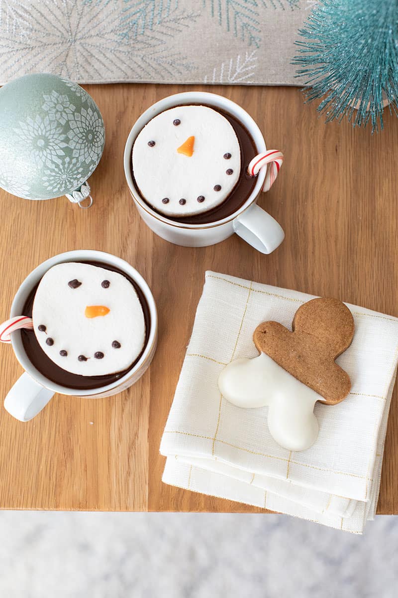 Table top with hot chocolate and snowmen marshmallows, a gingerbread cookie on a white and gold napkin and a sage ornament 