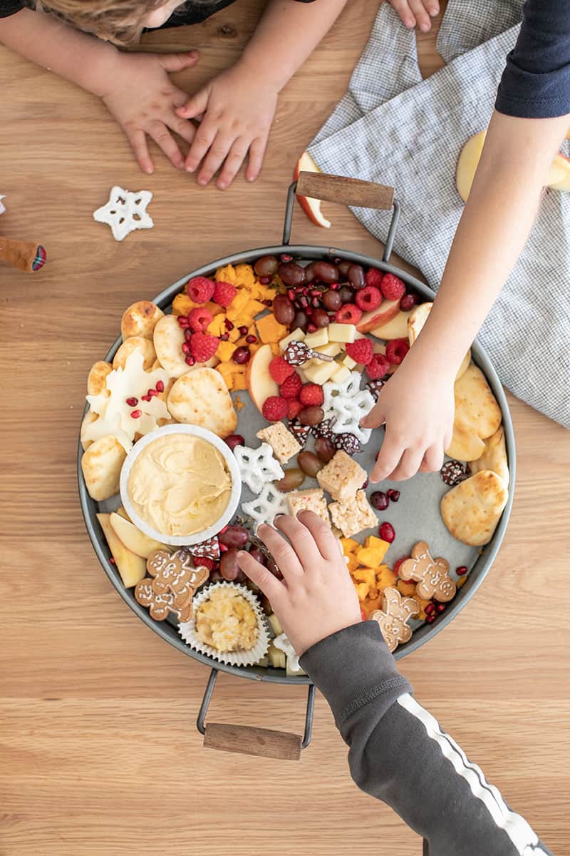 Kids reaching in to grab food from a round galvanized tray filled with hummus, cheese, pita, apples, yogurt covered pretzels and gingerbread cookies.