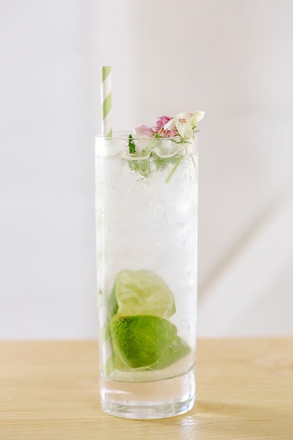 A classic gin and tonic in a highball glass with edible flowers, limes and a green paper straw.