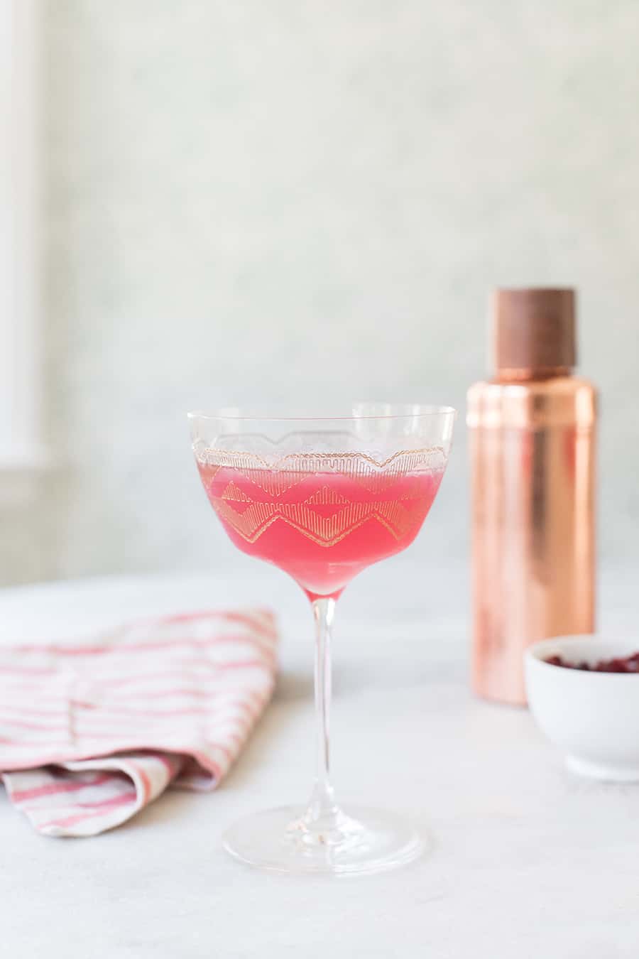 A pink cranberry martini in a gold coupe glass with a copper cocktail shaker. 