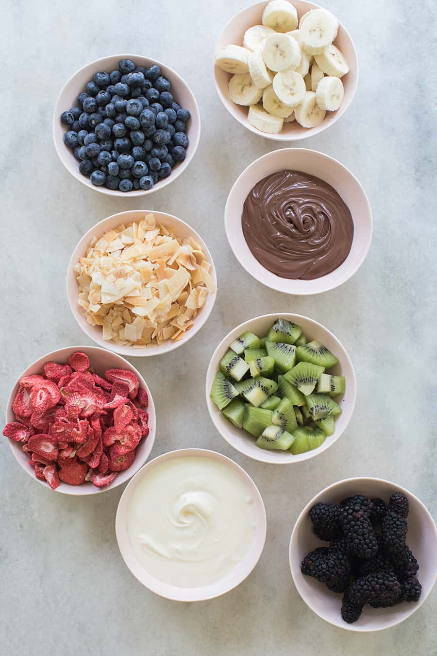 Kiwis, bananas, Nutella, blueberries and coconut shreds on pink bowls on the table.