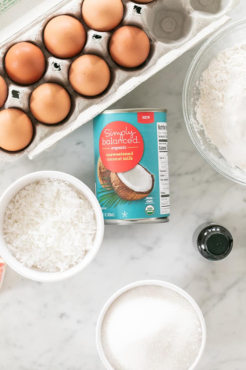 Canned coconut milk, eggs, shredded coconut and flour on a marble table.