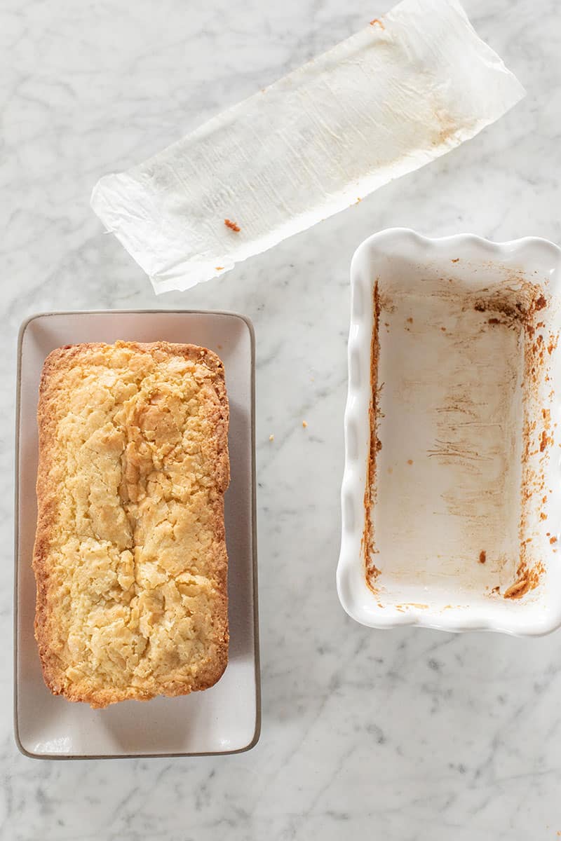 loaf cake on a platter, an empty loaf pan and a piece of parchment paper.