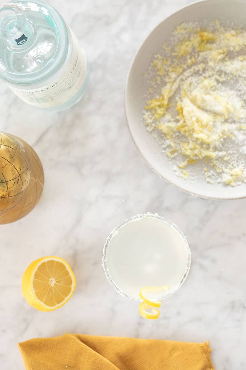 Lemon drop martini on a marble table with a yellow napkin, cocktail shaker and a bowl of lemon sugar.