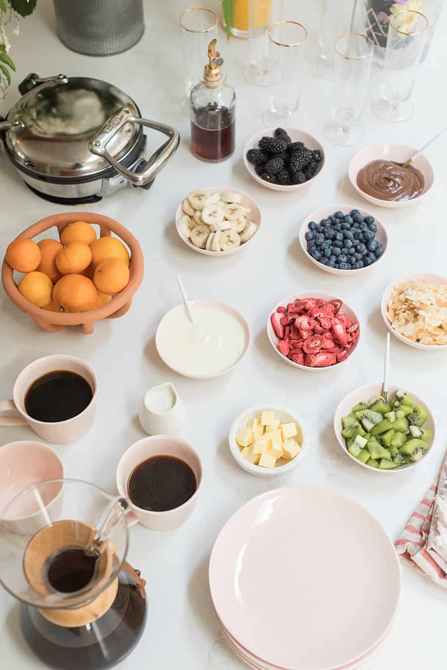 Waffle bar and toppings, mugs filled with coffee and a basket of oranges.