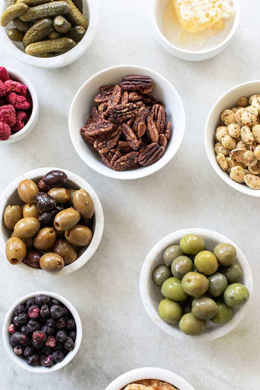 Olives, candied walnuts, almonds, dried blueberries in mini bowls on a marble table.