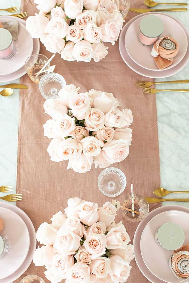 A pink table setting with roses and a pink table runner with pink dishes and gold flatware.