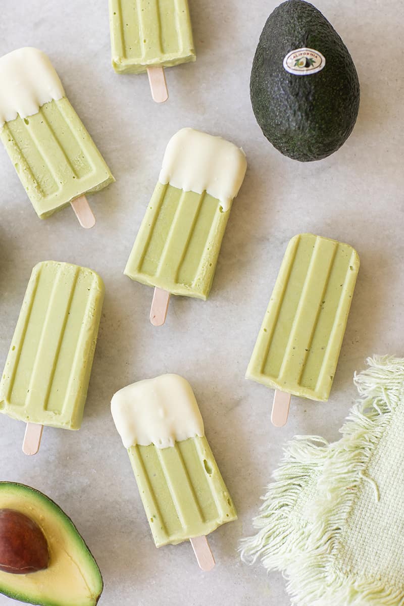 Avocado popsicles, dipped in white chocolate on a marble table with an avocado. 