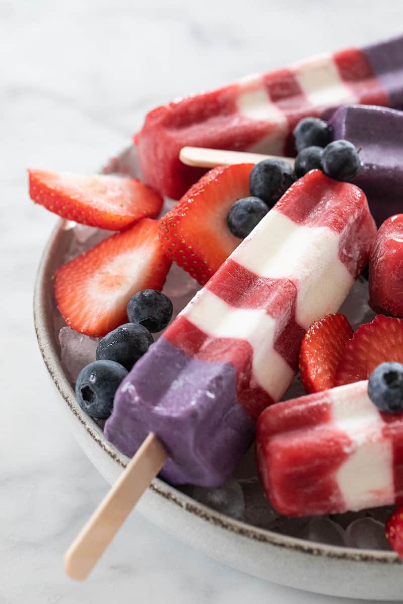 Striped red white and blue popsicle with strawberries, blueberries on a bowl of ice.