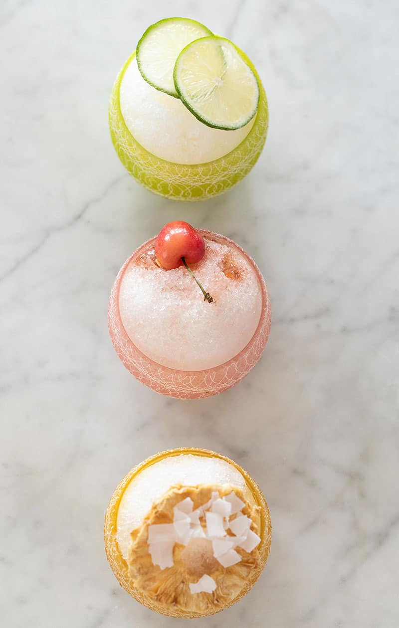 Three shaved ice in colorful cups with toppings.