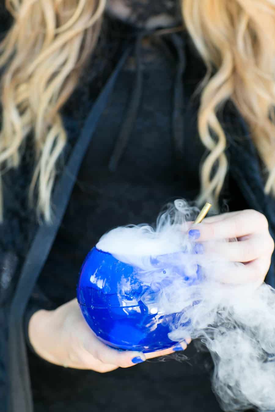 Girl holding a smoking blue cocktail with blue nails and a black cape. 