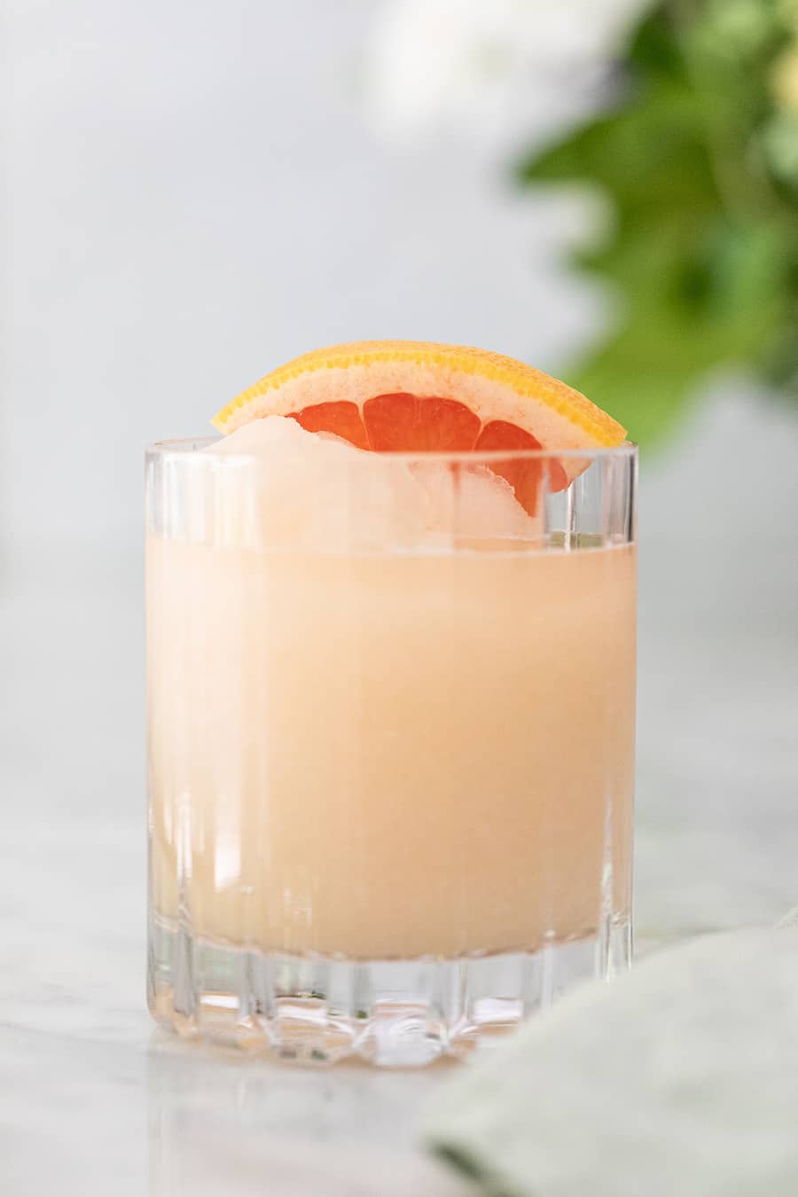 addison: A Paloma cocktail with large clear ice cubes in a tall collins  glass garnished with a grapefruit peel on a white marble counter top