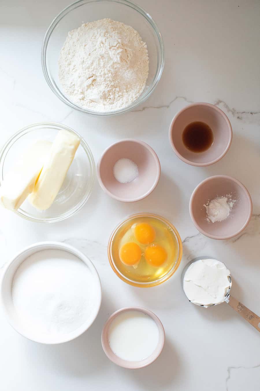 Flour, butter, eggs, vanilla, baking powder, salt, sour cream and sugar in bowls on a table.