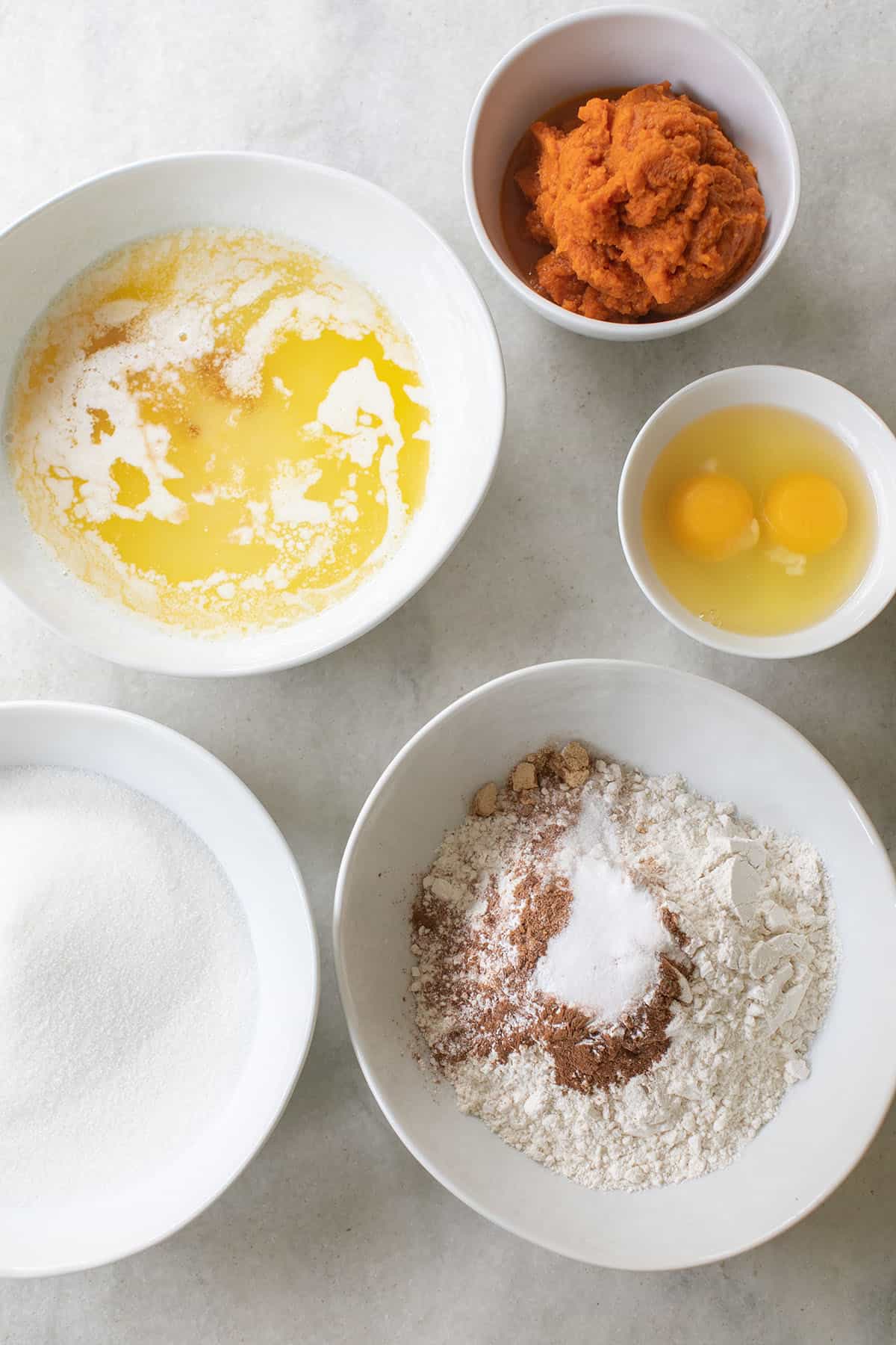 butter, eggs, pumpkin, sugar, flour, spices in white bowls on a marble table.
