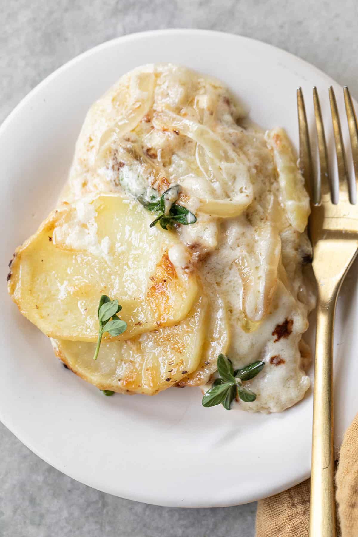 Scalloped potatoes on a plate with fresh thyme and a gold fork.