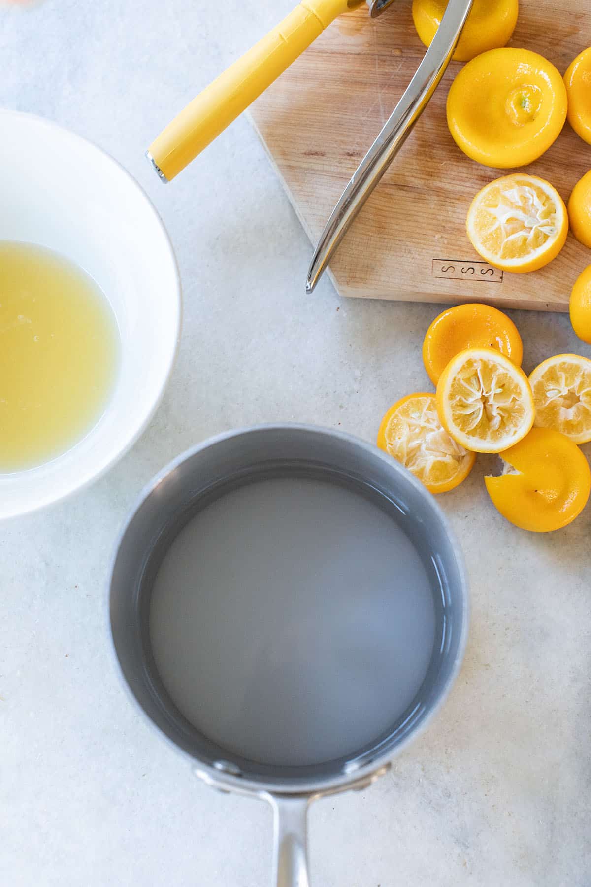 small saucepan filled with sugar and water with Meyer lemons on a table and a bowl of lemon juice. 