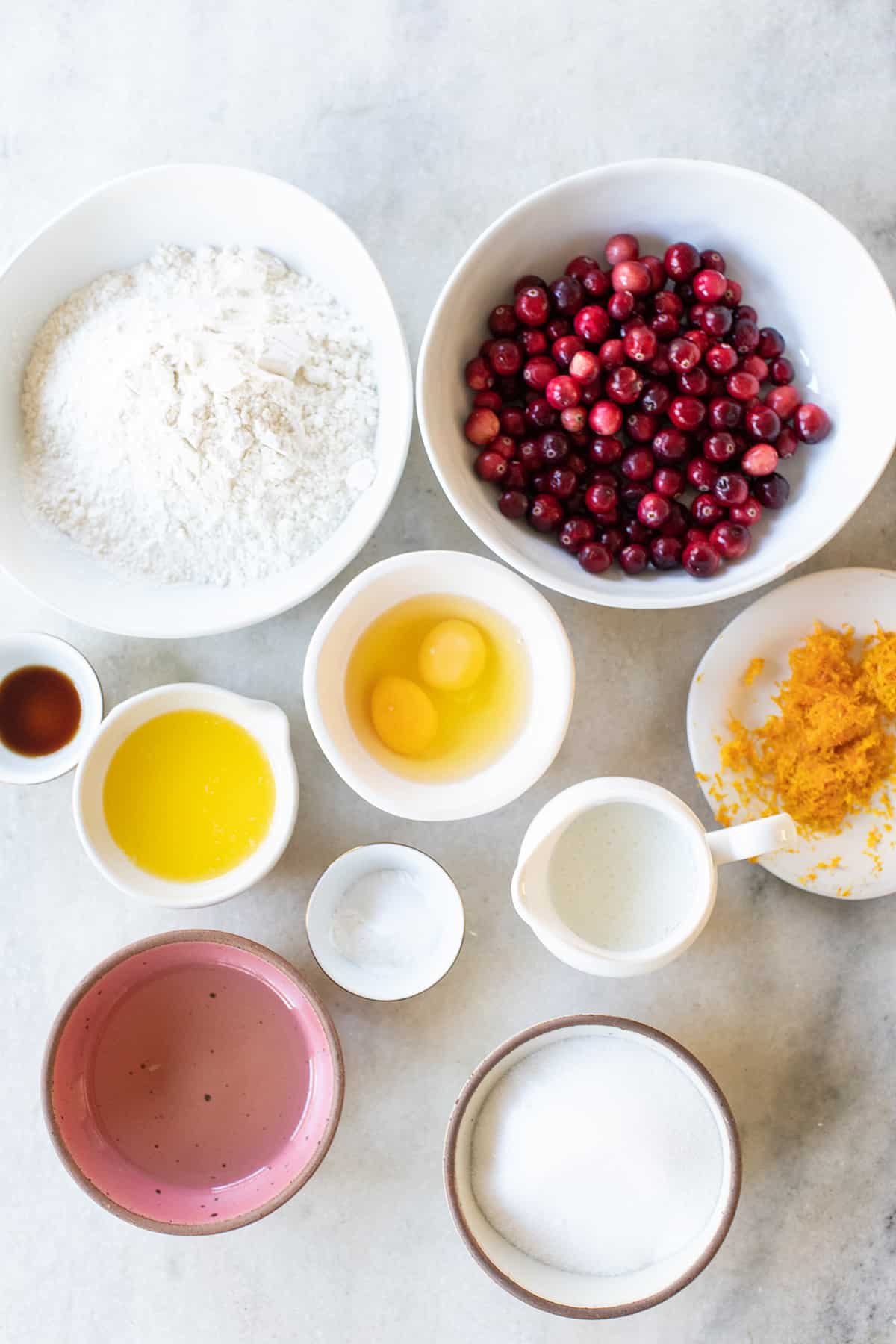 Flour, butter, sugar, eggs, orange zest, oil, fresh cranberries in bowls on a marble table.