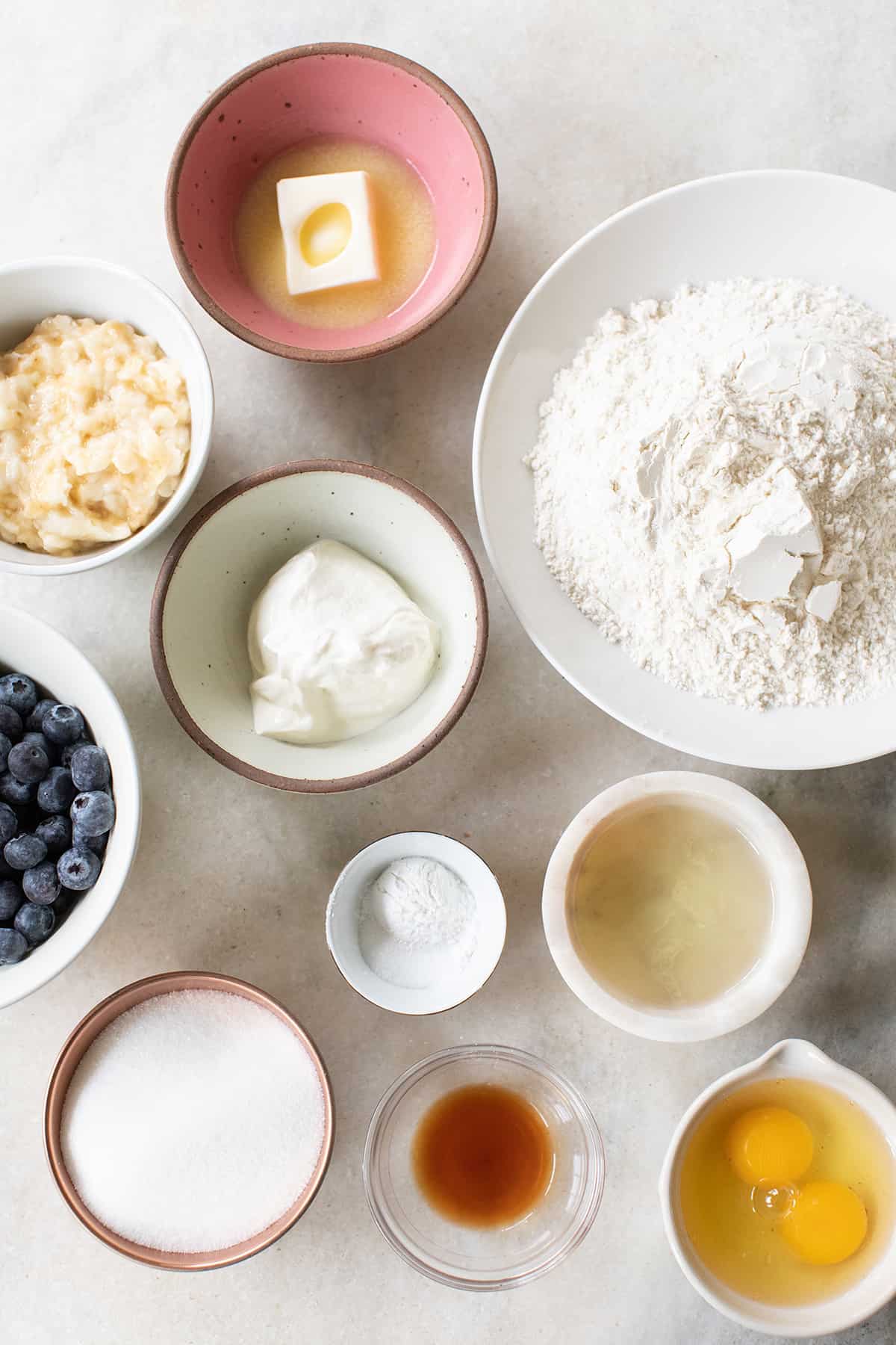 Eggs, flour, butter, sugar, vanilla, oil, sour cream, and blueberries in small bowls.