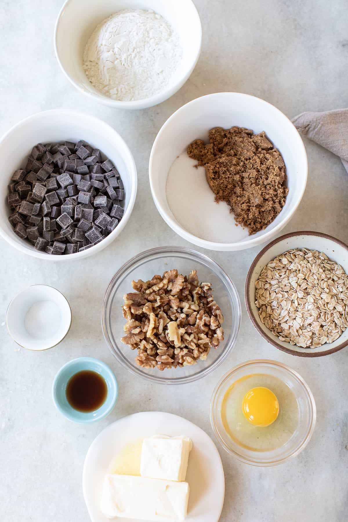 Brown sugar, butter, eggs, flour, walnuts, oats, vanilla extract, chocolate chunks in bowls on a marble table. 