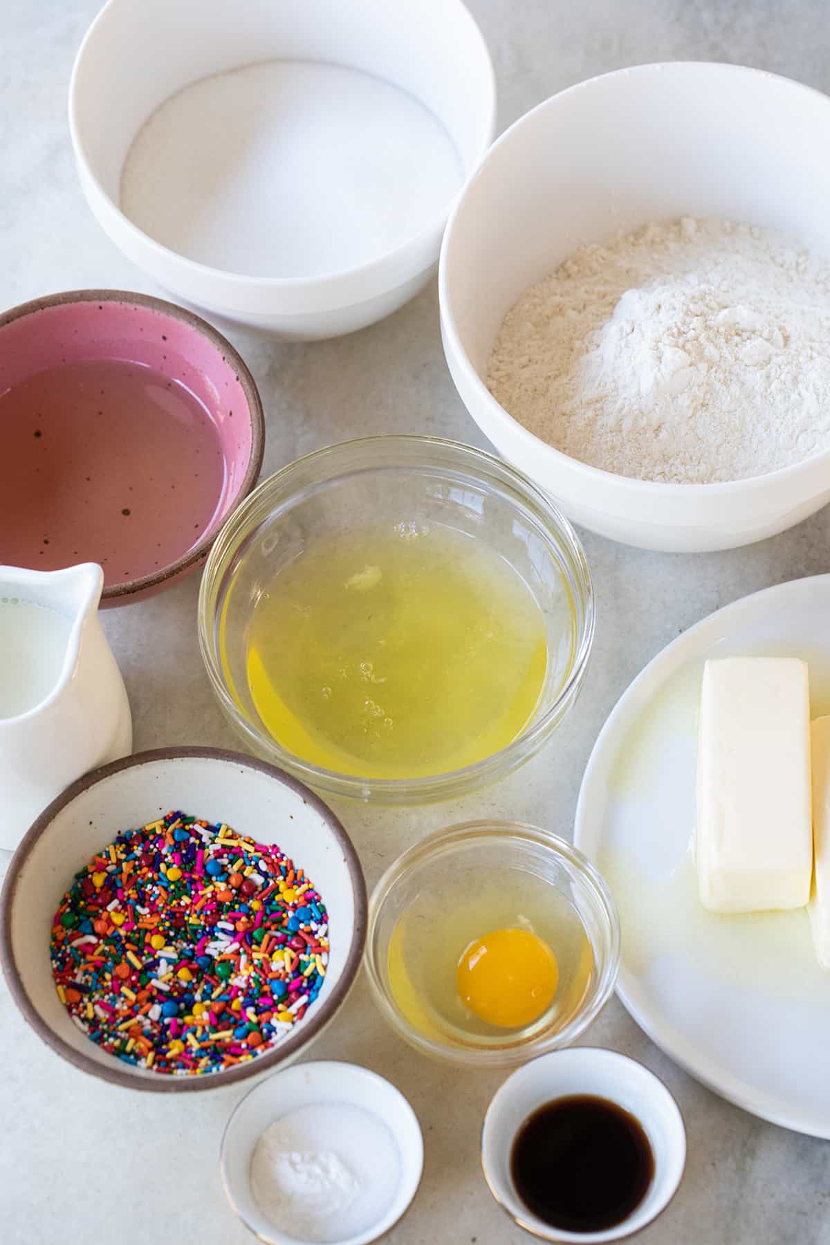 Oil, eggs, sugar, vanilla extract, flour, butter, milk and sprinkles in small bowls.