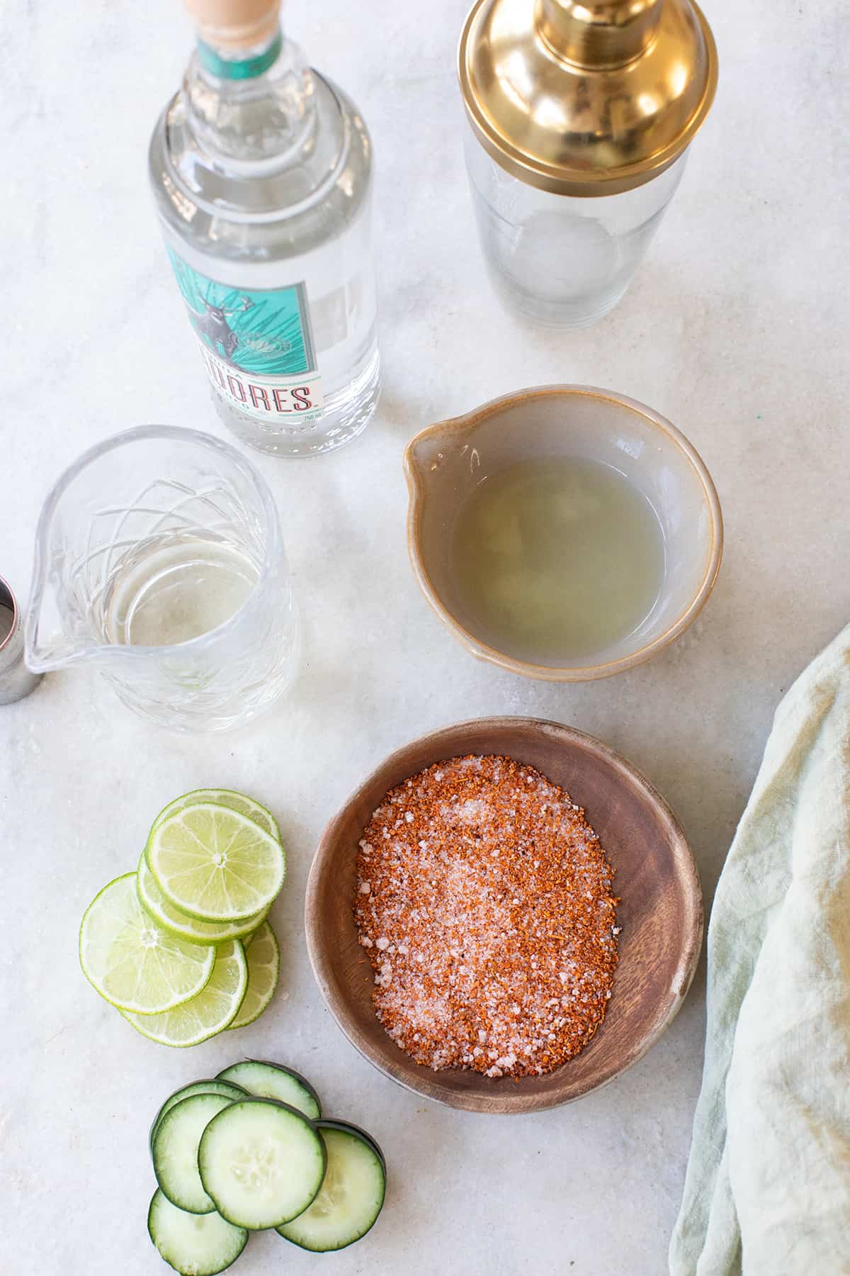 Lime juice, cucumbers, Tajín, tequila and a cocktail shaker on a table.