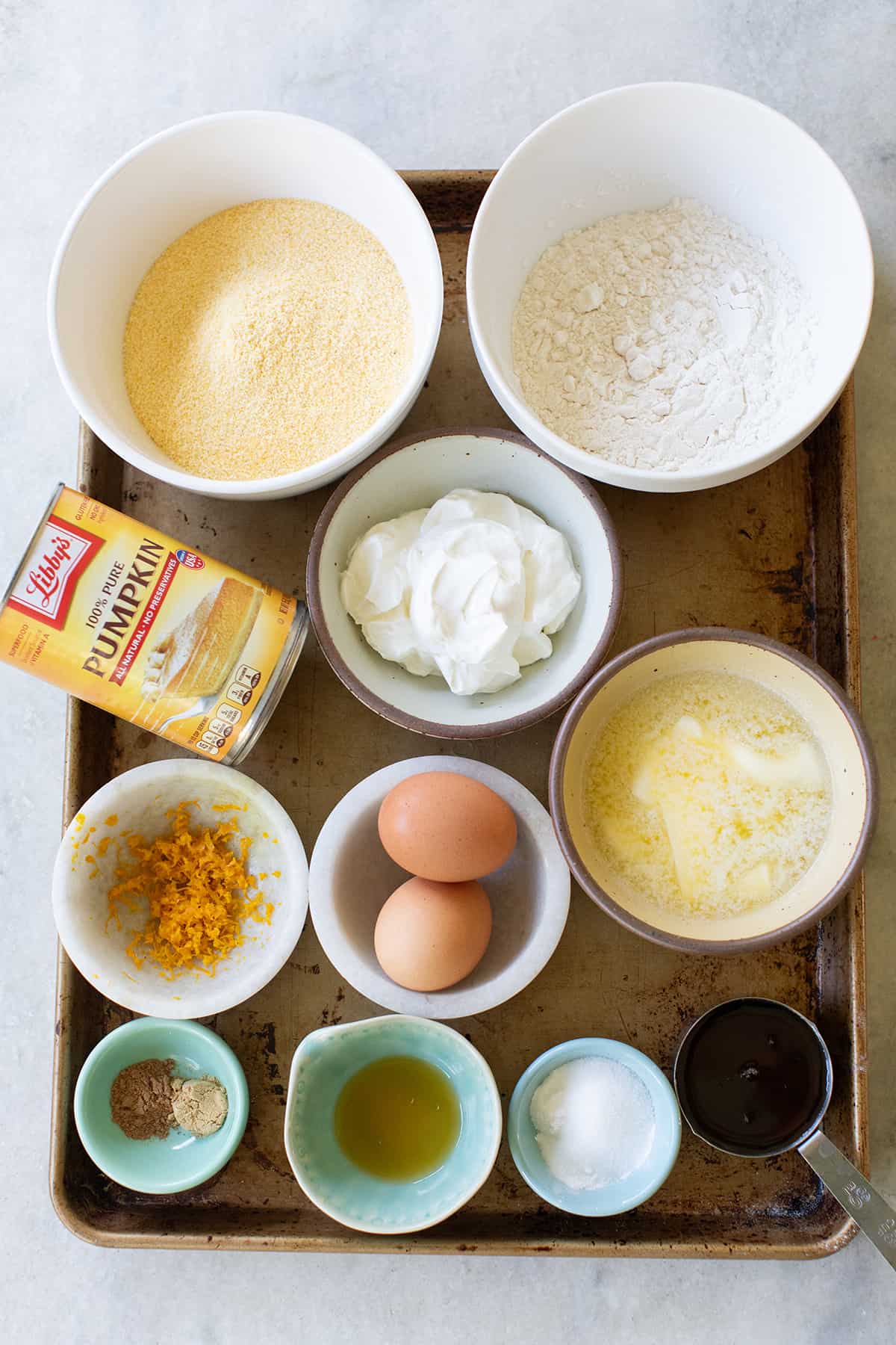 Cornmeal, pumpkin, flour, eggs, ground spices, sour cream, orange zest and maple syrup in bowls.