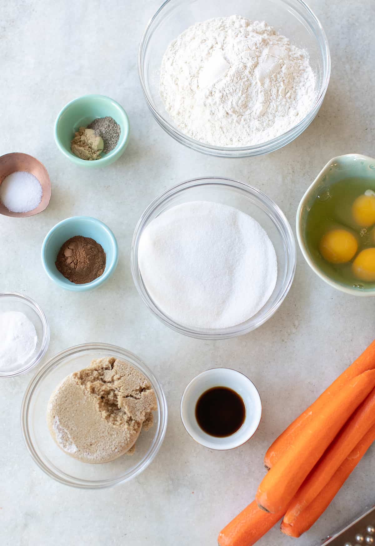 Carrots, flour, sugar, salt, eggs, vanilla and spices in small bowls to make a cake.