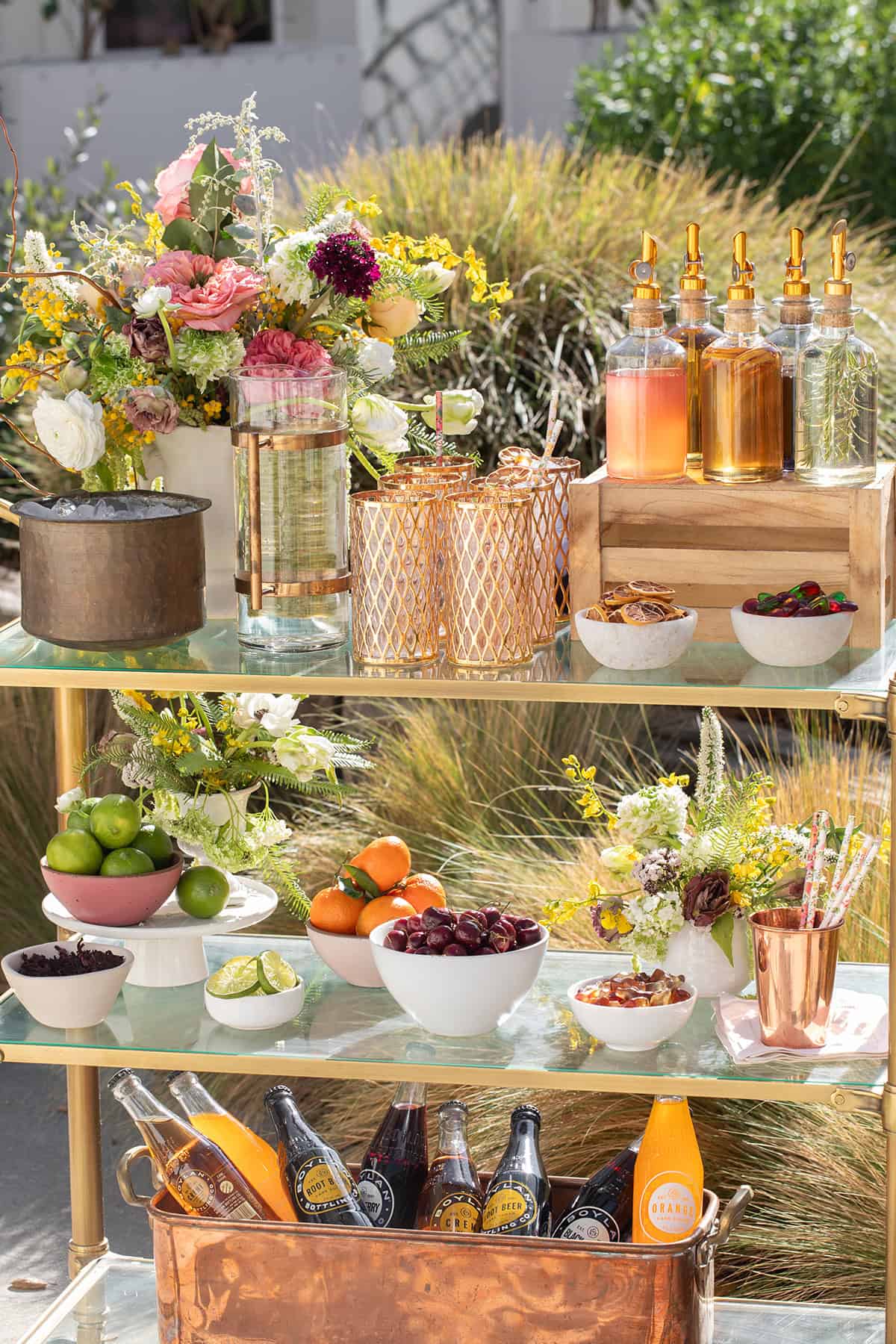 A soda bar set-up on a bar cart with flowers, syrups, glasses, and more.