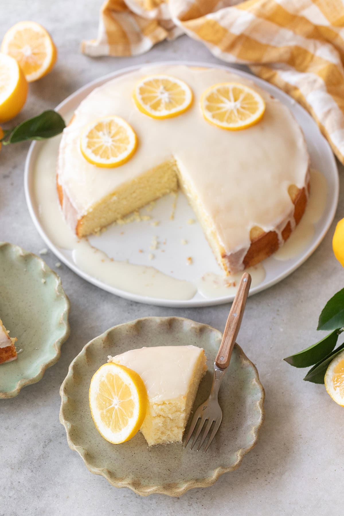 Lemon olive oil cake on table with slice removed green plates yellow napkins and fresh lemons.