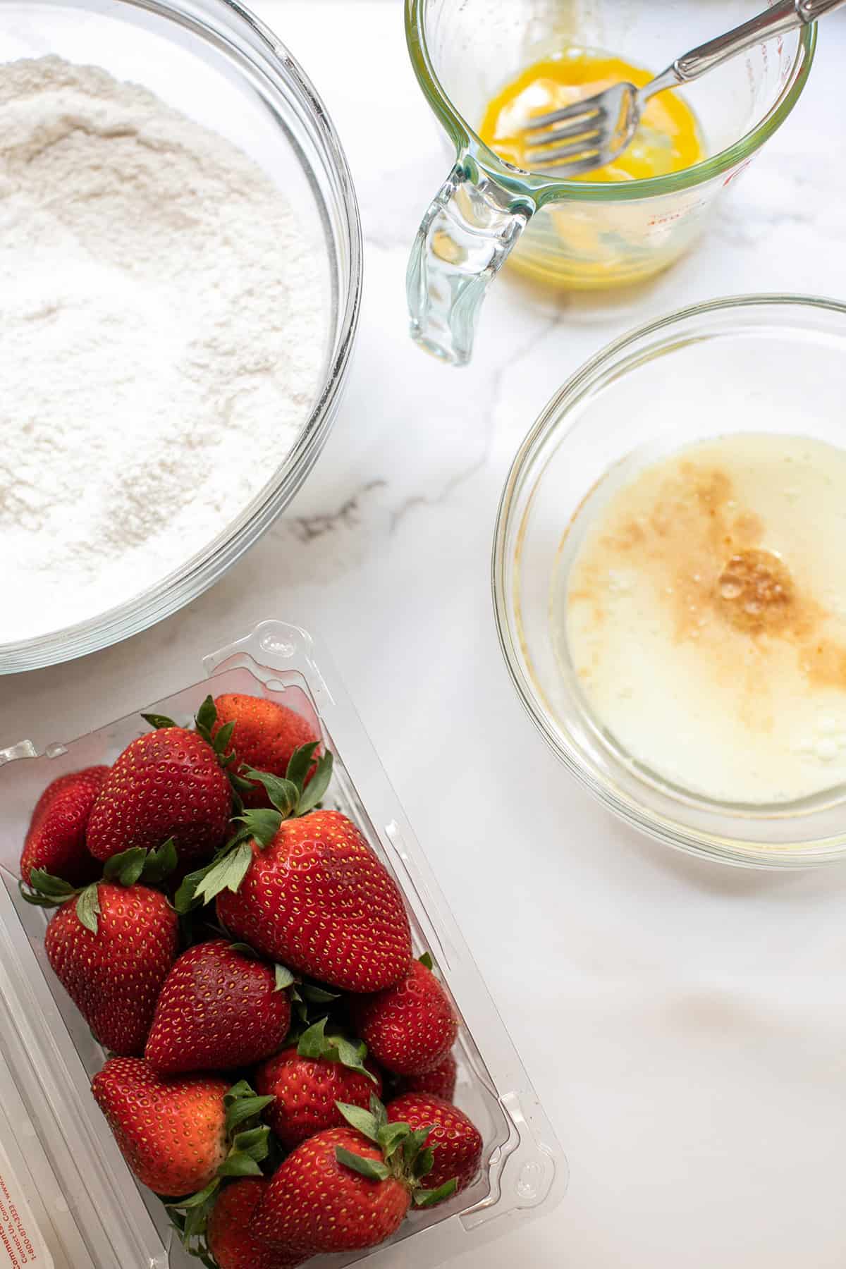 Ingredients for strawberry muffins including fresh strawberries, flour, sugar, eggs, and milk.