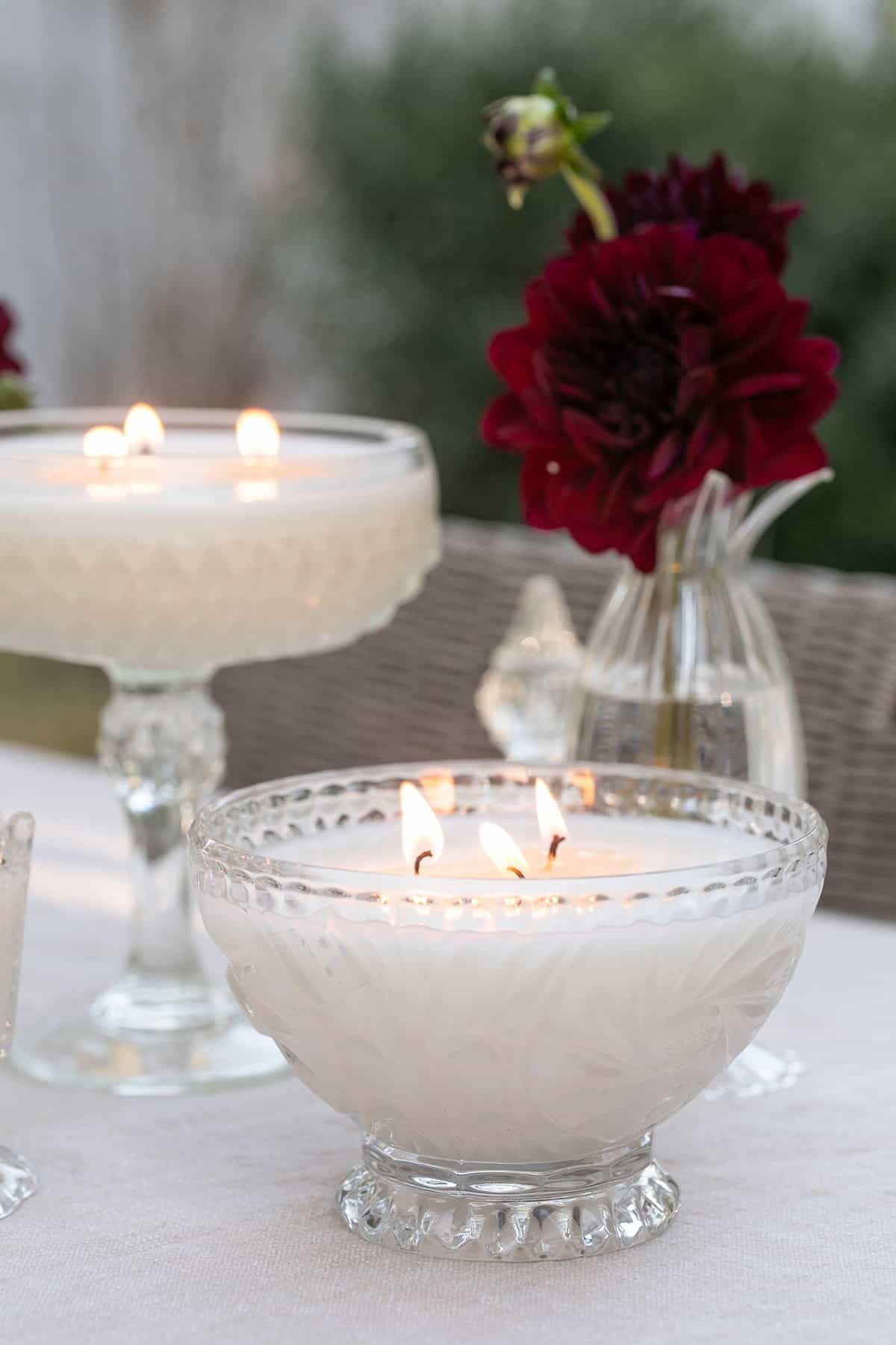 Homemade candle poured in a vintage glass vessel, lit on a table with flowers.